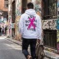 A man walking down a graffiti-covered city alley viewed from behind, wearing a white hoodie with black text reading 'ANTI SOCIAL SOCIAL CLUB' and a large pink X over the back.