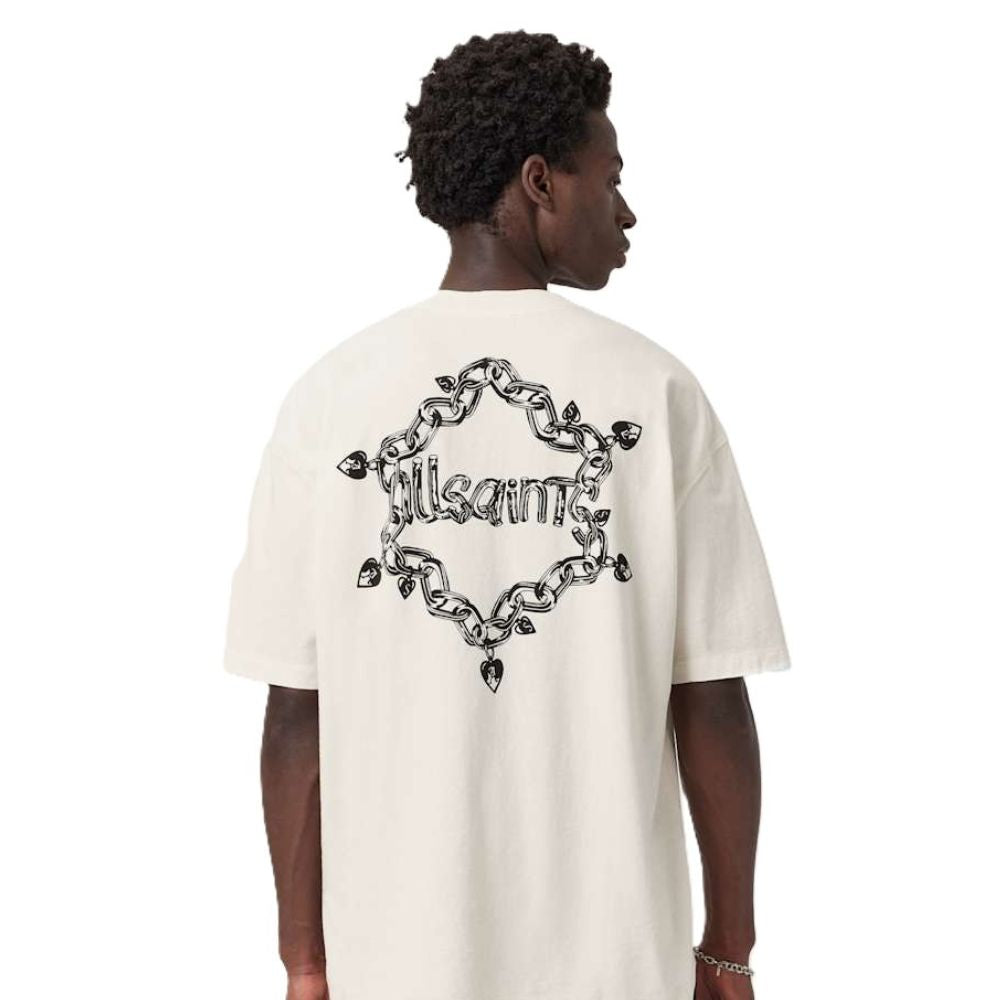 A man shown from the back wearing a cream-colored oversized t-shirt with a large black graphic on the back. The graphic features a chain-link heart design with the word 'Kith' in the center.