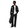 A model wearing a black knit cardigan with white graphic skull and cross designs, styled with a white t-shirt, dark jeans, black sunglasses, and carrying a black tote bag against a plain white background.