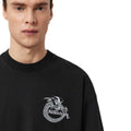 A close-up of a model wearing a black crewneck sweatshirt with a white graphic of a dragon wrapped in chains and the text 'All Saints' on the chest.