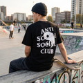 A man wearing a black beanie and a black t-shirt with a large white 'ANTI SOCIAL SOCIAL CLUB' yin-yang graphic on the back, sitting on a concrete ledge at a skatepark with a skateboard.