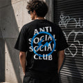 A man viewed from behind wearing a black t-shirt with a large graphic on the back. The graphic features the text 'ANTI SOCIAL SOCIAL CLUB' in white, overlaid with a blue circle and slash symbol.