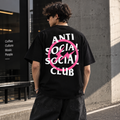 A man wearing a black t-shirt with a large graphic on the back, standing outdoors next to a concrete wall. The graphic features the text 'ANTI SOCIAL SOCIAL CLUB' in white, crossed out by a pink circle with a lightning bolt inside.