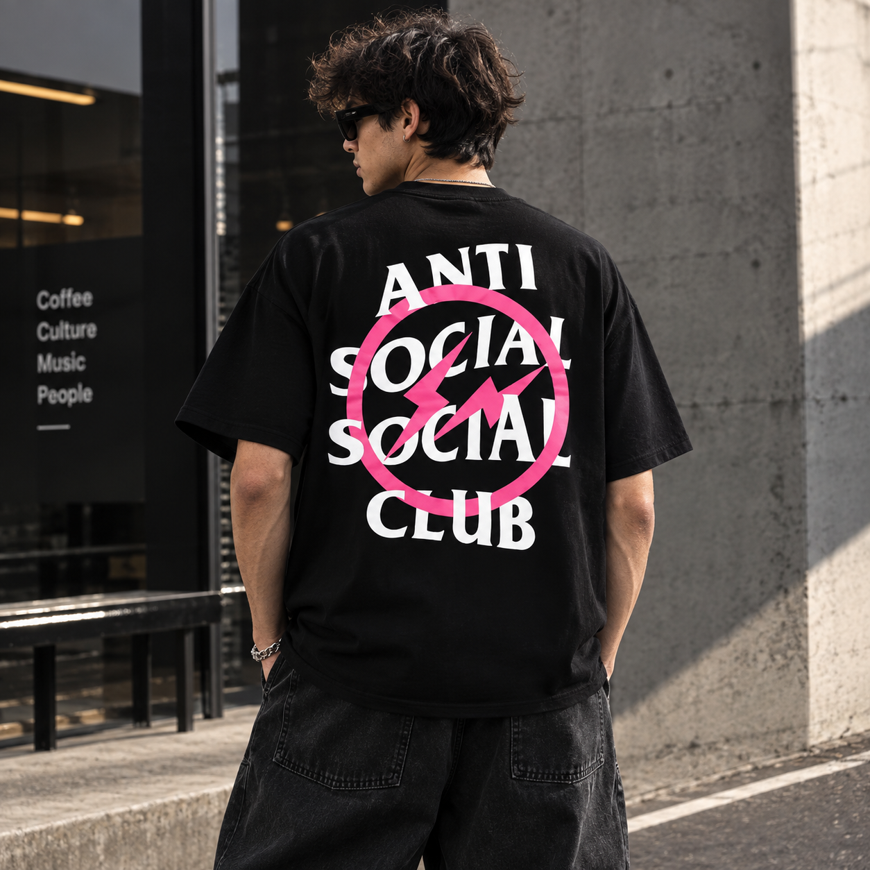 A man wearing a black t-shirt with a large graphic on the back, standing outdoors next to a concrete wall. The graphic features the text 'ANTI SOCIAL SOCIAL CLUB' in white, crossed out by a pink circle with a lightning bolt inside.
