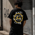 A person standing outdoors in front of a concrete wall and metal shutter, wearing a black T-shirt with a large back graphic that reads 'ANTI SOCIAL SOCIAL CLUB' in white text over a yellow circle and lightning bolt.