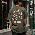 A man viewed from behind wearing an olive green t-shirt with 'ANTI SOCIAL SOCIAL CLUB' printed in pink text on the back, standing on a city street.