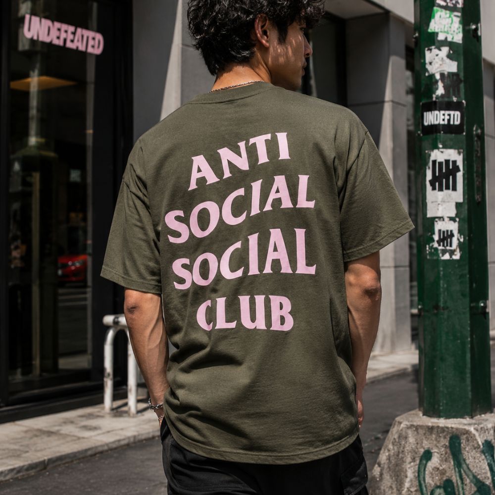 A man viewed from behind wearing an olive green t-shirt with 'ANTI SOCIAL SOCIAL CLUB' printed in pink text on the back, standing on a city street.