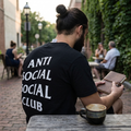 A man sitting at an outdoor cafe table, viewed from behind, wearing a black t-shirt with 'ANTI SOCIAL SOCIAL CLUB' printed in bold white letters on the back. He is holding a brown leather notebook, with a ceramic coffee mug on the wooden table in the foreground.