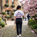 A woman viewed from behind walking on a cobblestone street, wearing a white graphic T-shirt with the words 'ANTI SOCIAL SOCIAL' in black and pink floral artwork, dark jeans, and carrying a woven straw bag.