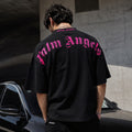 A man viewed from behind wearing a black Palm Angels t-shirt with a large pink gothic logo printed across the back, standing outdoors next to a black car in front of a concrete wall.