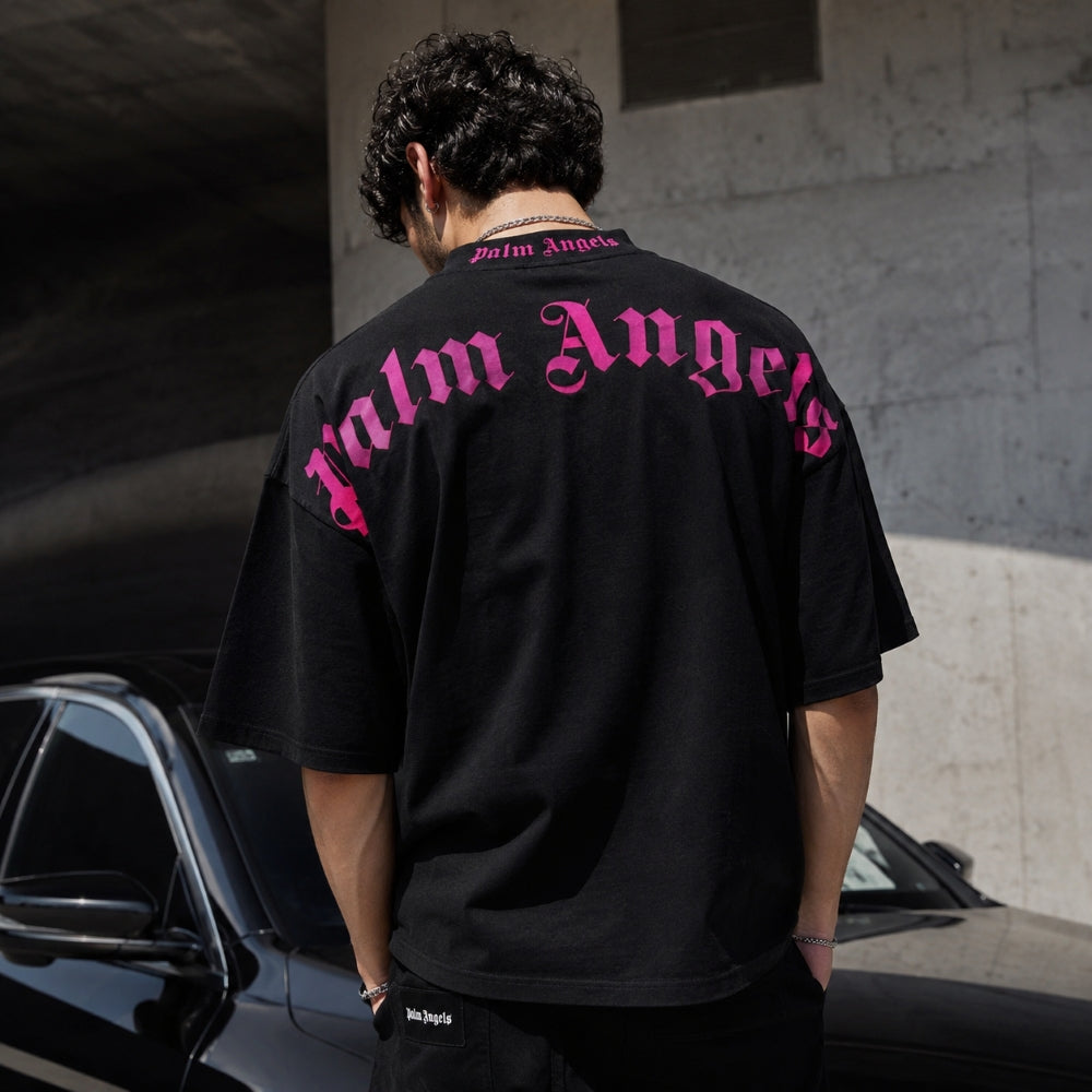 A man viewed from behind wearing a black Palm Angels t-shirt with a large pink gothic logo printed across the back, standing outdoors next to a black car in front of a concrete wall.