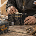 A person wearing a dark Carhartt jacket inserts cash into a camouflage bifold wallet with a transparent ID window, set on a rustic wooden workbench with work gloves and a Carhartt box nearby.