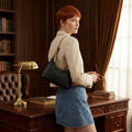 A woman with short red hair stands in a vintage-style library or study, wearing a black leather shoulder bag with a gold chain strap. She is dressed in a beige blazer and a blue denim skirt, posing next to a dark wood desk with a brass lamp and books.