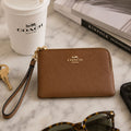 A brown pebbled leather Coach zip-around wallet with a wrist strap and gold logo, styled on a marble surface with a Coach takeaway cup, keys, a book, and sunglasses nearby.