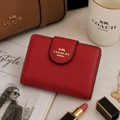 A red textured leather Coach wallet with a gold logo, styled on a tabletop with a brown Coach bag, a white Coach mug, lipstick, and other lifestyle props.