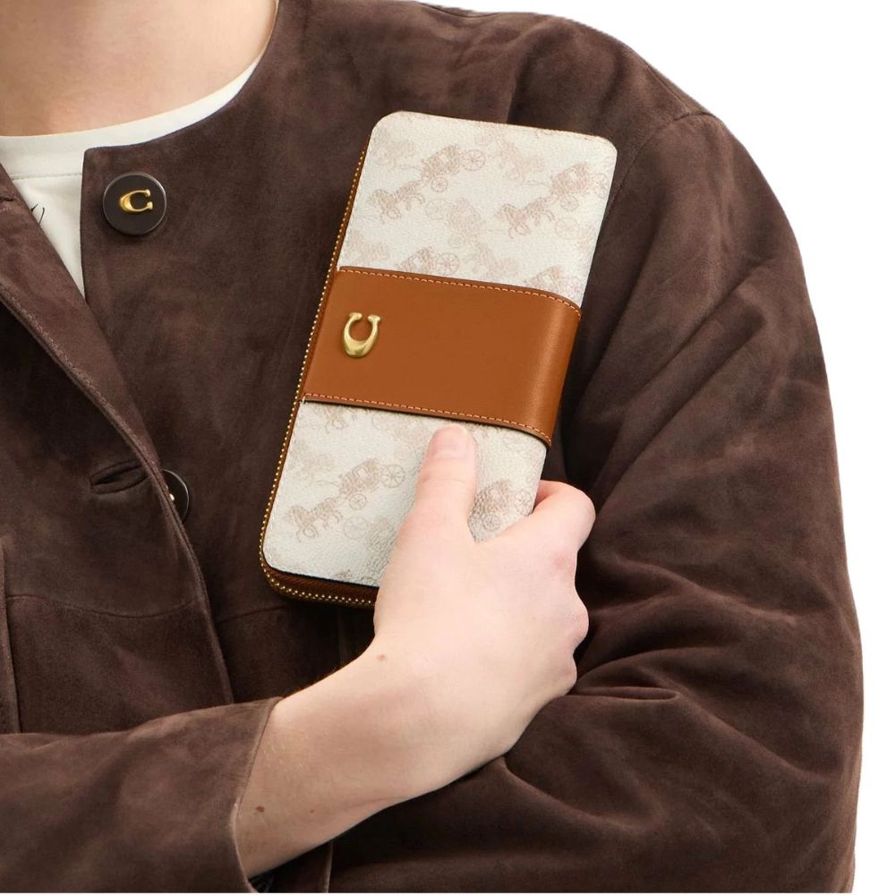 A close-up of a person wearing a brown suede jacket and holding a long wallet or notebook cover. The product has a cream-colored horse patterned body with a medium brown leather strap and a small gold horseshoe-shaped metal accent.