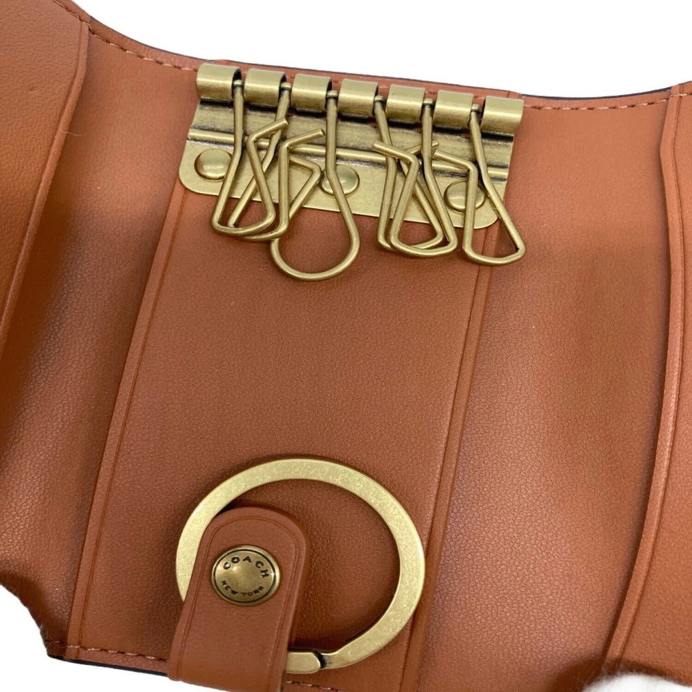 Close-up of a Coach leather bag featuring a warm tan-brown leather body, gold-tone metal hardware, a large circular ring detail, and a strap with four safety-pin style clips.