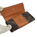 A Louis Vuitton monogram canvas and tan leather wallet held open by a white gloved hand, showing its interior card slots and snap-button closure on a white background.