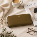 An olive green Coach zip-around wallet styled in a cozy flat-lay arrangement with a white mug of coffee, a magazine, eyeglasses, and a cream throw blanket.