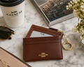A brown leather Coach card holder wallet with a gold-tone keychain ring, styled on a white marble surface with a Coach-branded coffee cup, sunglasses, a magazine, and flowers.