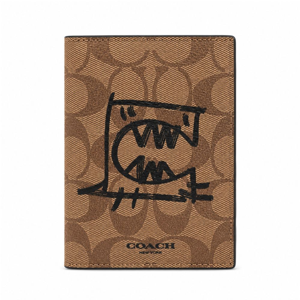 A Coach passport cover in brown signature canvas with a black hand-drawn graphic of a face inside a passport-stamp shape, and the Coach logo printed at the bottom.