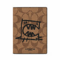 A Coach passport cover in brown signature canvas with a black hand-drawn graphic of a face inside a passport-stamp shape, and the Coach logo printed at the bottom.
