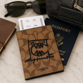 A Coach passport holder in the signature brown monogram canvas with a black graphic print, shown with a passport, boarding pass, sunglasses, notebook, and a Coach zip case on a light marble surface.