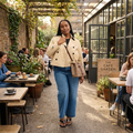 A woman walks through an outdoor cafe patio wearing a beige double-breasted blazer, blue jeans, and brown sandals, carrying a brown shoulder bag. The background shows other cafe patrons and a sign that reads 'THE CAFE GARDEN'.