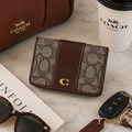 A Coach small bifold wallet with the brand's signature monogram canvas and brown leather trim, styled on a marble surface with a Coach bag, coffee cup, sunglasses, and keys nearby.
