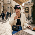 A woman sitting at a cafe table in a European street, wearing a cream ribbed cardigan, black top, blue jeans, and a black quilted shoulder bag with a gold C logo. She has a pink bow and green leaf hair clip.
