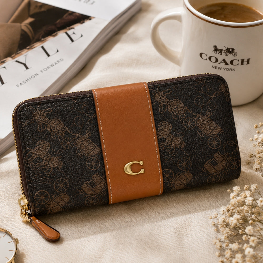 A Coach zip-around wallet in dark brown monogram canvas with a tan leather center stripe and a gold C logo, styled on a beige fabric surface next to a Coach coffee mug and a magazine.