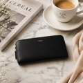A black leather zip-around wallet with gold-tone hardware and a small gold COACH logo, styled on a white marble surface with a coffee cup, magazine, and scarf nearby.