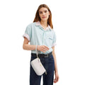 A model wearing a light blue and white striped short-sleeve button-up shirt, dark blue jeans, and a light gray crossbody bag, photographed against a solid white background.