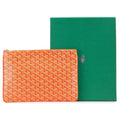 An orange Goyardine pouch with a repeating monogram pattern, displayed next to its green presentation box on a white background.