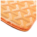 Close-up of an orange coated canvas or leather item featuring the signature Goyardine monogram pattern in white and dark brown, with a tan leather edge visible along the bottom left corner.