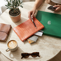 An orange textured zip pouch with a geometric pattern lies on a round beige table, surrounded by a green branded box, a brown leather notebook, a cup of latte, sunglasses, cards, and a phone. A person's hands are visible interacting with the scene.