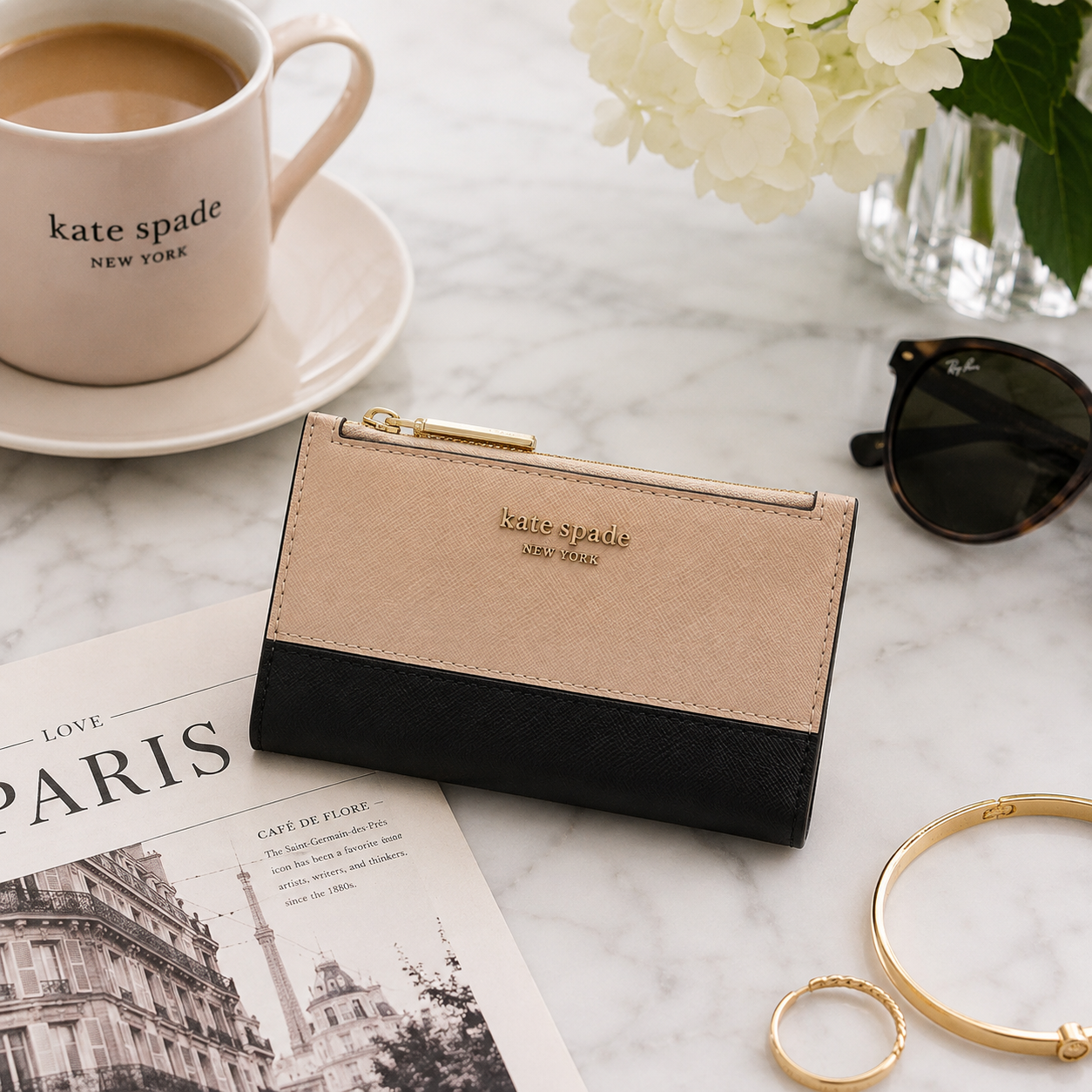 A Kate Spade New York two-tone wallet in beige and black leather, styled on a marble surface with a coffee cup, sunglasses, rings, and a magazine.