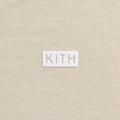 Close-up of a beige knit fabric with a small white rectangular patch centered on the chest featuring the word KITH in beige lettering.