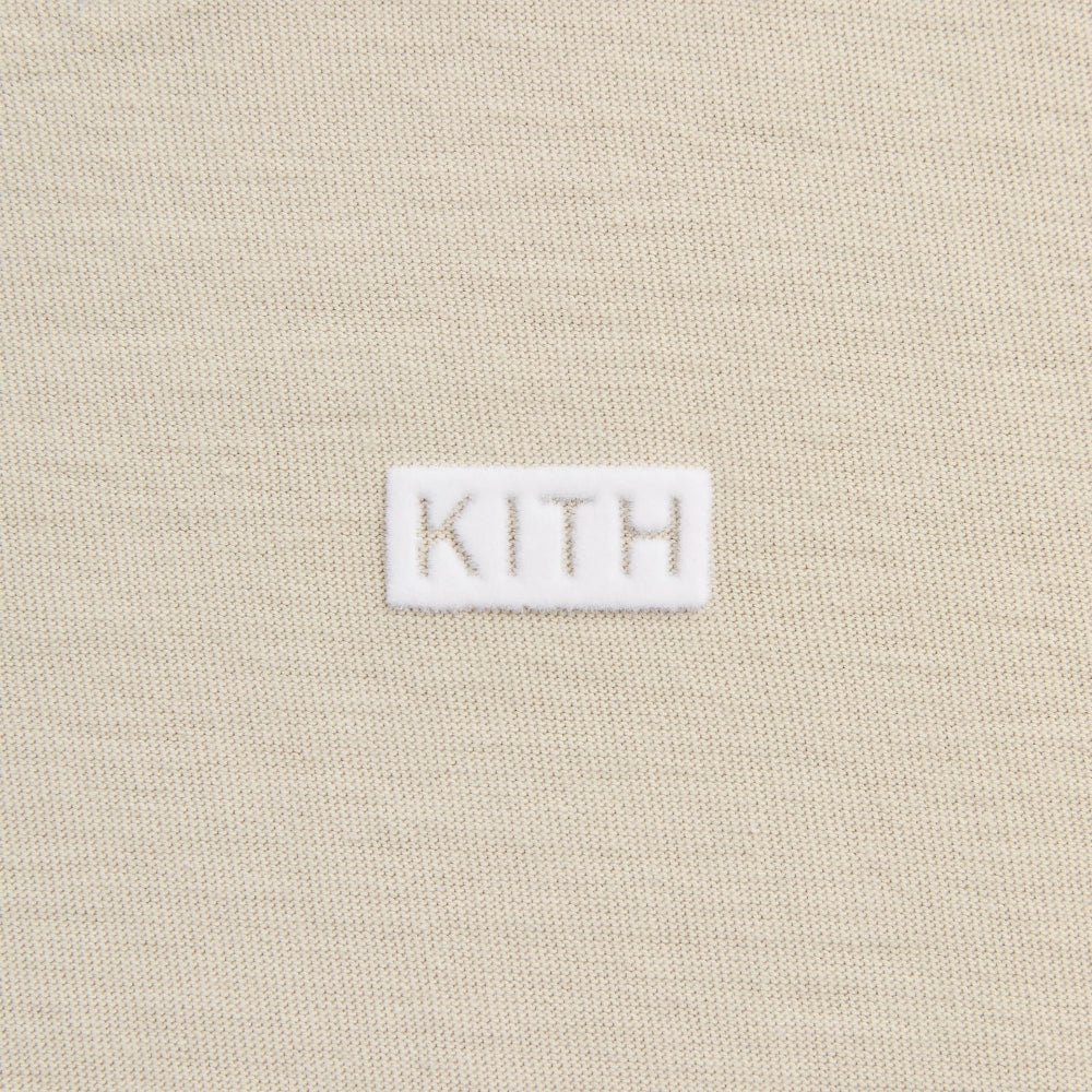 Close-up of a beige knit fabric with a small white rectangular patch centered on the chest featuring the word KITH in beige lettering.