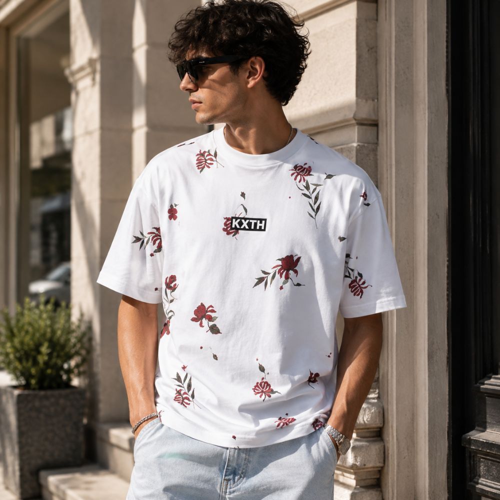A white short-sleeve crewneck t-shirt by KXTH, featuring a scattered red and green floral print and a black rectangular logo patch on the chest with the brand name 'KXTH' in white.
