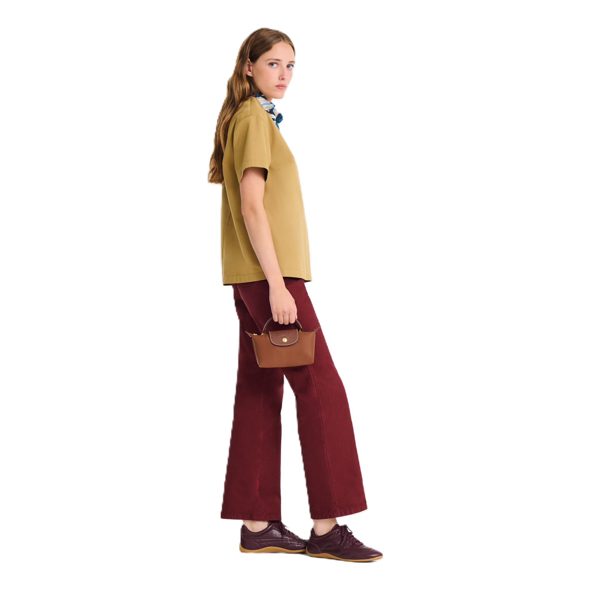 A woman stands in profile wearing a mustard yellow short-sleeve top, burgundy wide-leg pants, and dark brown shoes, holding a small brown handbag against a plain white background.