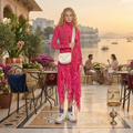 A woman stands on a luxurious outdoor terrace with a lake and palace in the background, wearing a vibrant pink patterned long-sleeve turtleneck dress with a white crossbody bag and white sneakers.