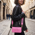 A woman in a black leather jacket carries a bright pink Marc Jacobs crossbody bag with gold hardware on a cobblestone street. The bag features a pink flap with the text 'THE MINI BAG MARC JACOBS' and a pink shoulder strap with 'COBS' printed on it.