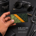 A hand holds a slim Michael Kors card holder wallet featuring a green monogram pattern and a diagonal mustard yellow stripe, set against a dark desk background with sunglasses, a coffee cup, a watch, and a notebook.