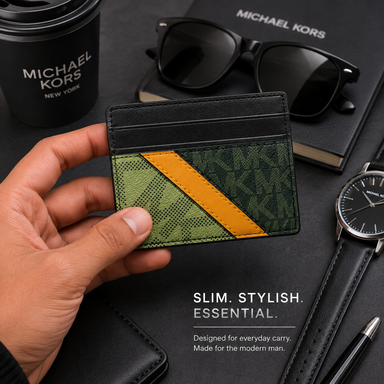 A hand holds a slim Michael Kors card holder wallet featuring a green monogram pattern and a diagonal mustard yellow stripe, set against a dark desk background with sunglasses, a coffee cup, a watch, and a notebook.