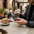 A man at a cafe holds a black leather cardholder wallet with a red accent stripe. The background shows a blurred outdoor cafe scene with people, a cup of coffee, and sunglasses on a table.