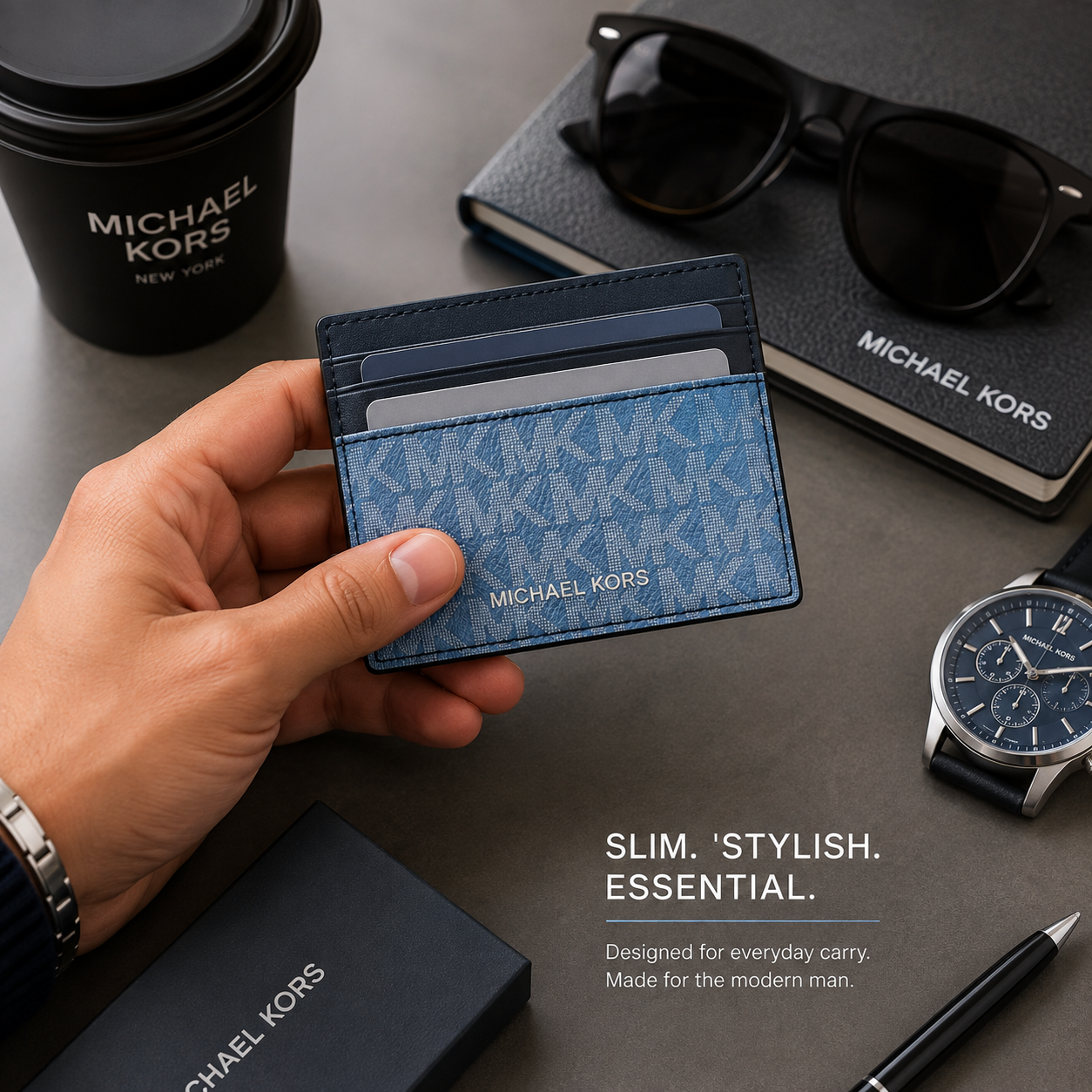 A hand holds a slim blue Michael Kors card holder wallet with a monogram pattern, surrounded by Michael Kors branded accessories including a coffee cup, notebook, sunglasses, watch, and box on a gray desk.