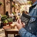 A person in a blue denim jacket holds a slim black cardholder wallet with a textured chevron pattern in an outdoor cafe setting.