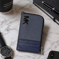 A textured zip-around wallet in a two-tone navy and blue-gray colorway, styled on a white marble surface with a watch, coffee cup, notebook, and smartphone nearby.