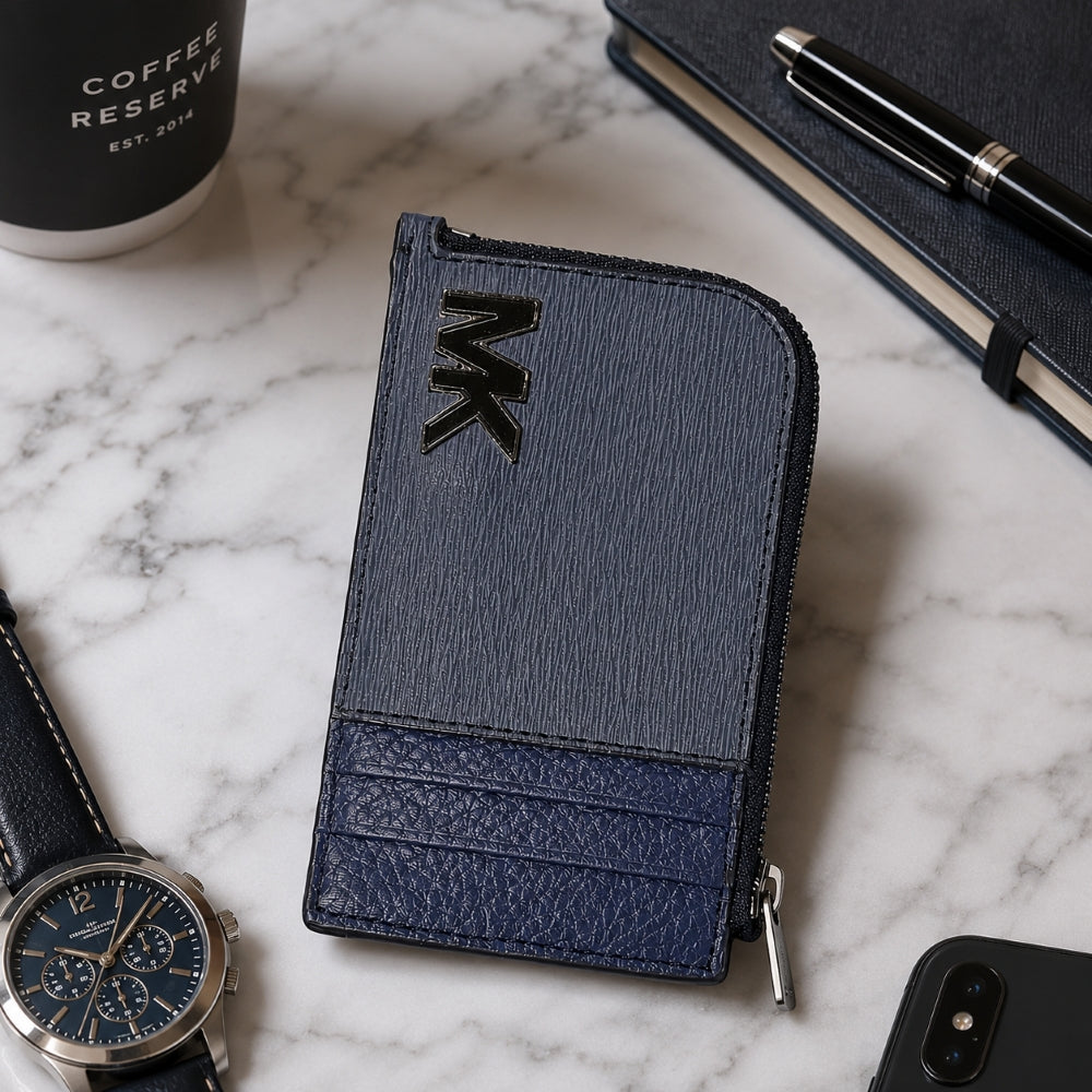 A textured zip-around wallet in a two-tone navy and blue-gray colorway, styled on a white marble surface with a watch, coffee cup, notebook, and smartphone nearby.